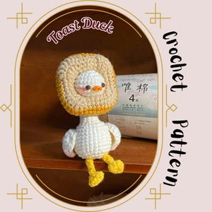 May include: A crocheted amigurumi toy duck wearing a crocheted toast hat. The duck is white with yellow feet and a yellow beak. The toast hat is brown and yellow. The duck is sitting on a wooden shelf.