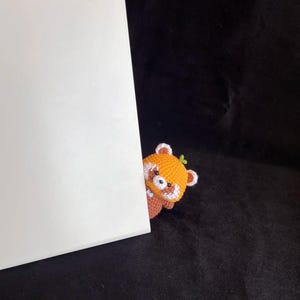 K&ouml;nnte beinhalten: Ein handgeh&auml;kelter roter Panda, der hinter einem wei&szlig;en rechteckigen Objekt hervorlugt. Der rote Panda hat orange-wei&szlig;es Fell mit kleinen braunen Ohren und einem gr&uuml;nen Blatt auf dem Kopf. Der Hintergrund ist schwarz.