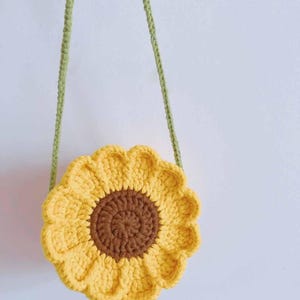 May include: A crocheted sunflower-shaped bag with a brown center and yellow petals. The bag has a green strap for carrying. The bag is handmade and has a whimsical, floral design. The bag is a unique accessory.