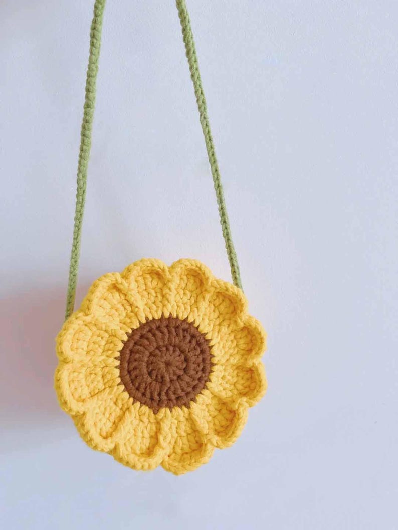 May include: A crocheted sunflower-shaped bag with a brown center and yellow petals. The bag has a green strap for carrying. The bag is handmade and has a whimsical, floral design. The bag is a unique accessory.