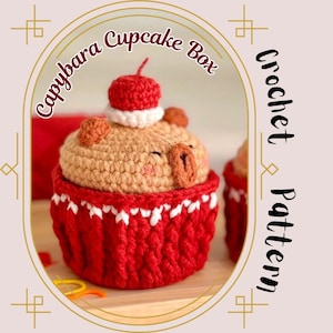 May include: A crocheted cupcake box in the shape of a capybara. The box is primarily red and tan, with a cherry on top. The text "Capybara Cupcake Box" and "Crochet Pattern" are visible.