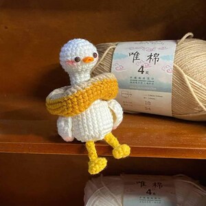 May include: A white crocheted goose wearing a yellow and brown crocheted life preserver. The goose is sitting on a wooden shelf with two balls of yarn in the foreground. The yarn is labeled "唯棉 4" in Chinese characters.
