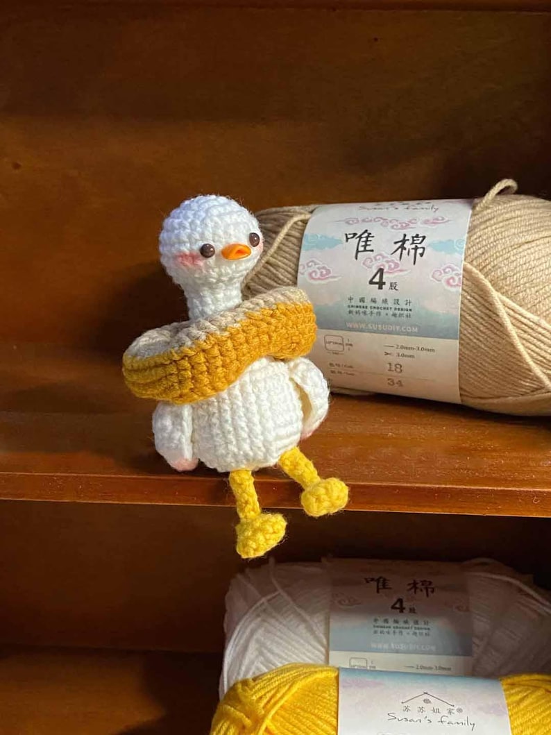 May include: A white crocheted goose wearing a yellow and brown crocheted life preserver. The goose is sitting on a wooden shelf with two balls of yarn in the foreground. The yarn is labeled "唯棉 4" in Chinese characters.