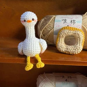 May include: A white crocheted goose with yellow feet and a brown yarn ball in the background. The yarn ball has a label that says "Peony 4".