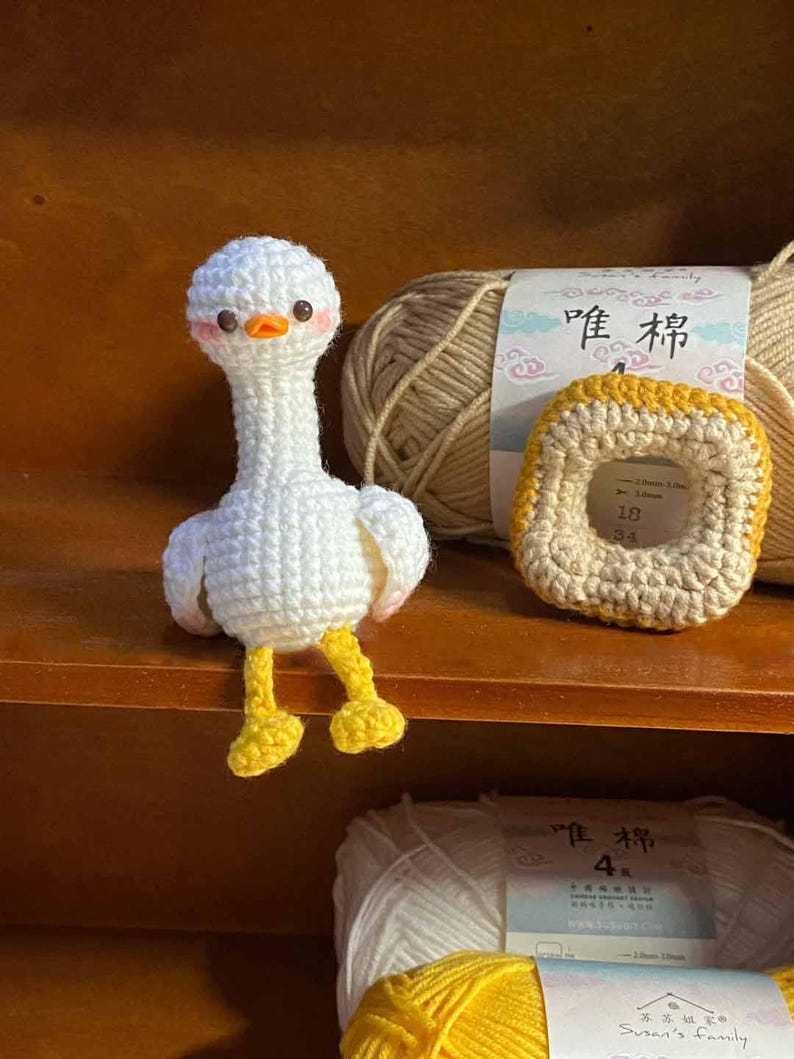 May include: A white crocheted goose with yellow feet and a brown yarn ball in the background. The yarn ball has a label that says "Peony 4".