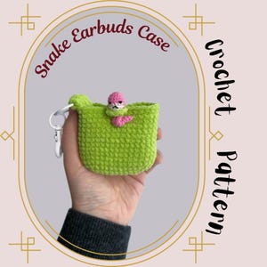May include: Crochet pattern for a green snake-shaped earbud case with a pink and white snake head. The case has a zipper and a key chain loop. "Snake Earbuds Case Crochet Pattern" is written on the image.