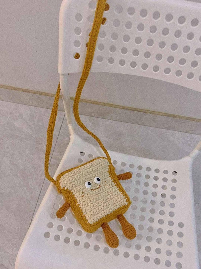 K&ouml;nnte beinhalten: Eine geh&auml;kelte Tasche in Brotform mit einem l&auml;chelnden Gesicht und braunen Armen und Beinen. Die Tasche h&auml;ngt an einem wei&szlig;en Stuhl.