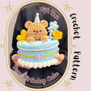 Op de afbeelding: Gehaakte berenverjaardagstaart met een lichtblauwe basis, gele accenten en een bruine berentopper. De taart is versierd met bloemen, sterren en hartvormige versieringen. De tekst op de afbeelding luidt "Gift Box" en "Crochet Pattern".