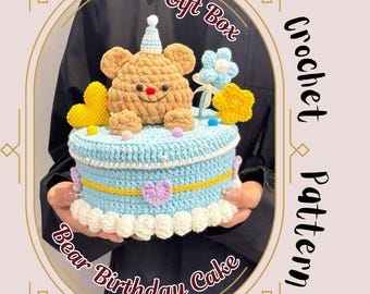 May include: Crocheted bear birthday cake with a light blue base, yellow accents, and a brown bear topper. The cake is decorated with a flower, star, and heart-shaped embellishments. Text on the image reads "Gift Box" and "Crochet Pattern".
