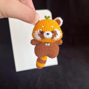 K&ouml;nnte beinhalten: Geh&auml;kelte Rotpanda-Figur. Das handgefertigte Tier hat einen braunen K&ouml;rper, einen orangefarbenen Kopf und Schwanz sowie wei&szlig;e Akzente. Es hat eine kleine blaue Fliege und ein gr&uuml;nes Blatt auf dem Kopf. Der Rotpanda wird vor einem wei&szlig;en Hintergrund gehalten.