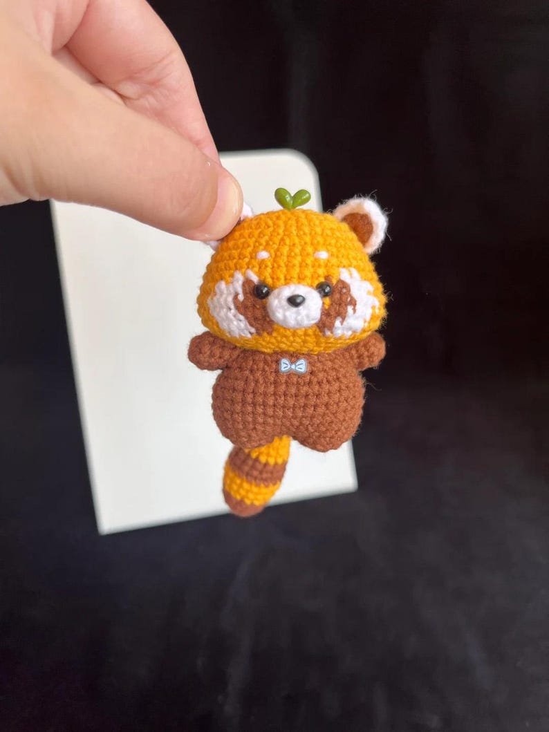 K&ouml;nnte beinhalten: Geh&auml;kelte Rotpanda-Figur. Das handgefertigte Tier hat einen braunen K&ouml;rper, einen orangefarbenen Kopf und Schwanz sowie wei&szlig;e Akzente. Es hat eine kleine blaue Fliege und ein gr&uuml;nes Blatt auf dem Kopf. Der Rotpanda wird vor einem wei&szlig;en Hintergrund gehalten.