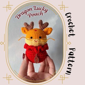 Puede incluir: Una bolsa de crochet con forma de dragón con cuerpo rojo, cabeza amarilla y cuernos marrones. La bolsa tiene un amuleto dorado con caracteres chinos. La bolsa está etiquetada como "Dragon Lucky Pouch Crochet Pattern".