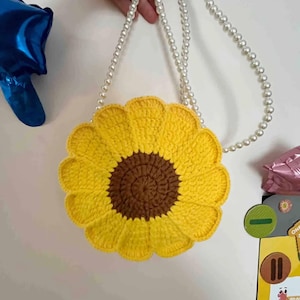 May include: A yellow crochet sunflower-shaped purse with a brown center and a pearl beaded strap. The bag is held up by a hand. The background is a plain white wall.