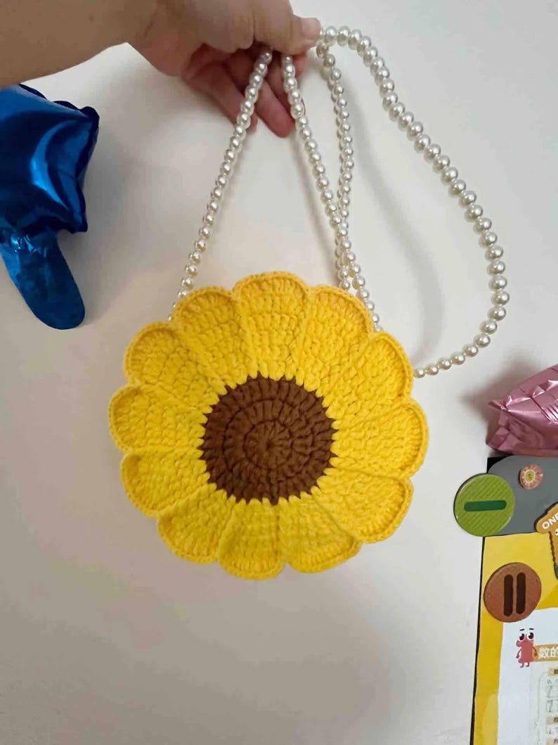 May include: A yellow crochet sunflower-shaped purse with a brown center and a pearl beaded strap. The bag is held up by a hand. The background is a plain white wall.