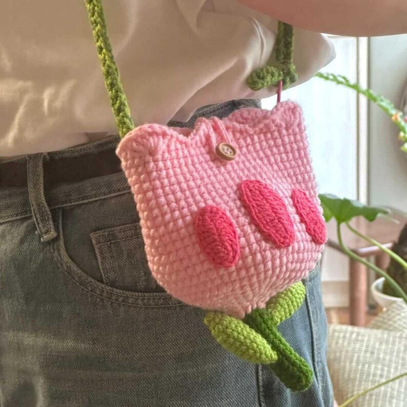 May include: A pink crocheted tulip-shaped bag with a green crocheted stem and leaves. The bag has a button closure and a green strap.