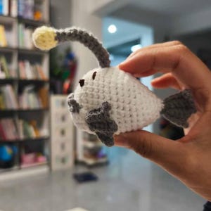 K&ouml;nnte beinhalten: Ein handgefertigtes geh&auml;keltes Anglerfisch-Spielzeug. Der Fisch ist wei&szlig; mit grauen Flossen und einer grauen, gebogenen Antenne mit einer gelben Kugel. Das Spielzeug wird in einer Hand gehalten, um die Details der H&auml;kelarbeit zu zeigen.