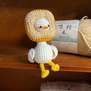 May include: A white crocheted amigurumi duck wearing a crocheted brown and yellow bread hat. The duck is sitting on a shelf with balls of yarn in the background.