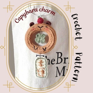 Könnte beinhalten: Ein gehäkelter Capybara-Anhänger mit einem Kleeblatt-Anhänger, der daran hängt. Der Anhänger ist braun mit einer roten Nase und weißen Augen. Der Text "Capybara charm" ist oben im Bild geschrieben. Der Text "Crochet Pattern" ist rechts im Bild geschrieben.