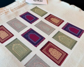 May include: Off-white tote bag with a repeating pattern of colorful prayer rug illustrations. The rugs feature intricate designs in shades of red, green, blue, and beige. A tag with the text "Crescent & Star" is attached.