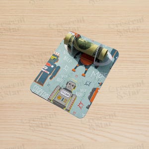 May include: A light blue money holder with a robot design. The holder has a rolled-up bill secured by a clear plastic band. The design features various robots, speech bubbles, and the word "SPACEBOTS".