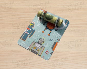 May include: A light blue money holder with a robot design. The holder has a rolled-up bill secured by a clear plastic band. The design features various robots, speech bubbles, and the word "SPACEBOTS".
