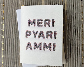 May include: A white card with the words "MERI PYARI AMMI" spelled out in colorful, glittery letters. The letters are made up of small, colorful beads.