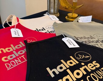 May include: A display of aprons in red, black, beige, and olive green. Each apron features the words "halal calories DON'T COUNT" in gold lettering, along with a halal symbol. The aprons are made of a sturdy fabric and have a neck strap.