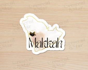 May include: A sticker featuring a watercolor map outline of Saudi Arabia with a gold border. The sticker includes a black cube and the word "Makkah" in a stylish font, with a watercolor wash background.
