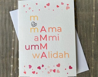 May include: A white greeting card with the words "mAma aMmi umM wAlidah" in various colors. The card has small pink and red hearts scattered around the top and bottom. The card is inside a white envelope.