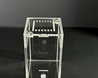May include: A clear acrylic enclosure featuring a rectangular vertical design with a top lid containing a series of small ventilation holes arranged in a square pattern. The base of the enclosure displays a small circular logo and white text.