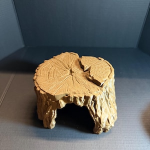 May include: A light brown, hollow tree stump replica featuring a textured bark exterior and a detailed cross-section top with visible growth rings. The object has a wide, arched opening at the base, creating a cave-like structure with a flat top.
