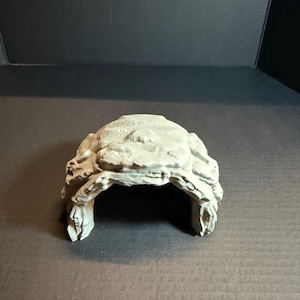 Könnte beinhalten: Eine hellgraue, 3D-gedruckte bogenförmige Struktur mit einer texturierten, geschichteten Oberfläche. Das Objekt verfügt über eine hohle Mitte und eine raue, unebene Oberseite, die natürlichen Felsformationen ähnelt.