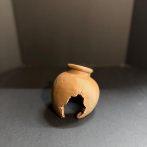 May include: A small, round, light brown clay pot with a narrow neck and a wide rim. The body of the vessel features a large, jagged, irregular opening on the lower side, revealing the hollow interior and the textured thickness of the clay material.