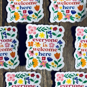 Pu&ograve; includere: Adesivi bianchi con la scritta "everyone is welcome here" circondati da disegni floreali colorati. Gli adesivi presentano vari fiori nei toni del rosa, arancione, giallo e blu, su uno sfondo di legno.