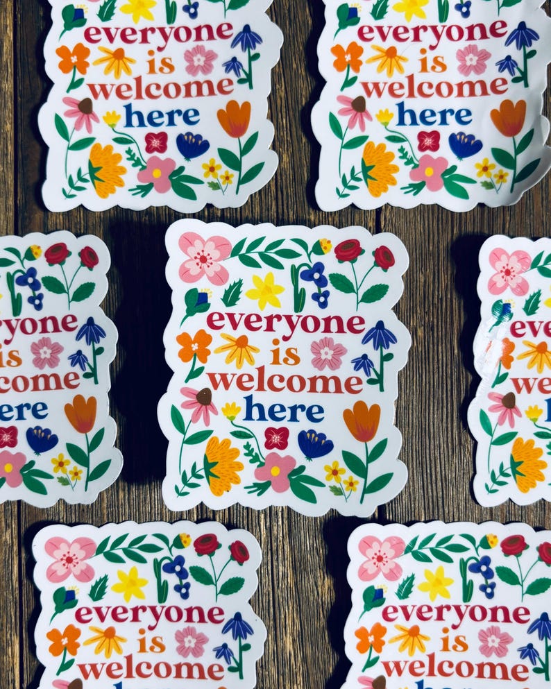 Pu&ograve; includere: Adesivi bianchi con la scritta "everyone is welcome here" circondati da disegni floreali colorati. Gli adesivi presentano vari fiori nei toni del rosa, arancione, giallo e blu, su uno sfondo di legno.