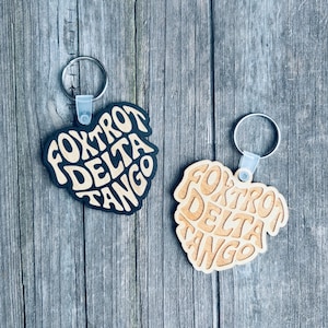 May include: Two keychains with the text "FOXTROT DELTA TANGO" in a stylized font. One keychain is black with cream-colored text, and the other is natural wood with a lighter brown text. Each keychain has a silver keyring.