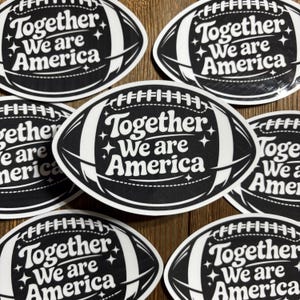 Può includere: Una collezione di adesivi neri e bianchi a forma di pallone da football. Ogni adesivo presenta il testo "Together We are America" in un font stilizzato con accenti di stelle. Gli adesivi sono disposti su una superficie di legno.
