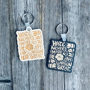 May include: Two keychains with the message "Hate Never Made Any Nation Great." One keychain is light beige with brown floral accents, the other is navy blue with beige accents. Both have silver key rings.