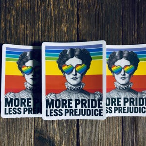 May include: Three stickers featuring a vintage illustration of a woman wearing heart-shaped rainbow sunglasses. The background is a rainbow gradient. The text "MORE PRIDE LESS PREJUDICE" is printed in black at the bottom.