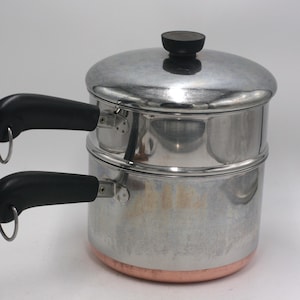 May include: A stainless steel double boiler with a copper-colored base. The pot features two black heat-resistant handles, each with a small metal hanging loop at the end, and a matching stainless steel lid with a black knob on top.