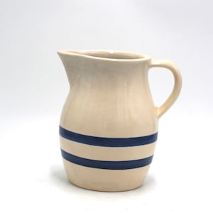May include: Cream-colored ceramic pitcher with a curved handle and spout. Two horizontal navy blue stripes encircle the body of the pitcher. The pitcher is a classic design, suitable for serving beverages.