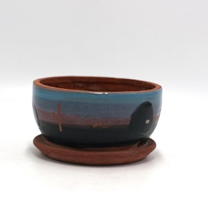 May include: A small terracotta planter with a blue, purple, and black glaze. The planter has a simple design with a few lines and a circle. It comes with a matching saucer.