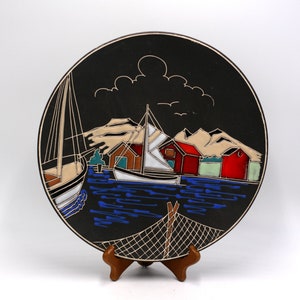 May include: A black decorative plate with a coastal scene. The plate features sailboats, red and white buildings, and a blue sea. The design includes a white outline and is displayed on a wooden stand.