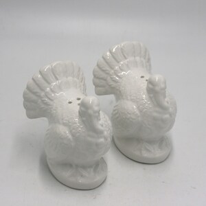 May include: A set of two white ceramic turkey salt and pepper shakers. The turkeys are facing each other with their tails up.