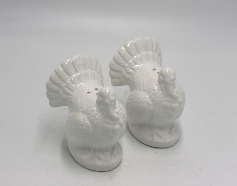 May include: A set of two white ceramic turkey salt and pepper shakers. The turkeys are facing each other with their tails up.