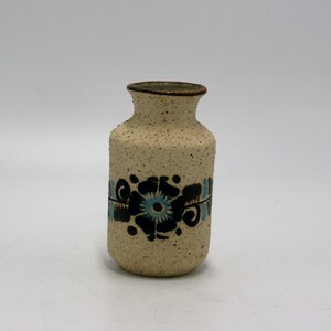 May include: A small, white ceramic vase with a speckled texture and a brown rim. It features a blue and black floral design around the middle.