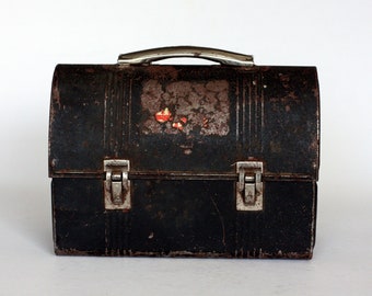 May include: A vintage metal lunchbox with a black finish and a silver handle. The lunchbox has a worn and faded appearance with some rust spots. It has two silver clasps on the front.
