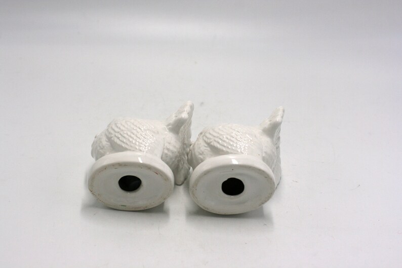 May include: Two white ceramic owl figurines with textured feathers. The owls are facing each other and have a hole in the bottom of their bases.