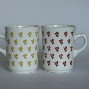 May include: Two white ceramic mugs with handles. One mug has a pattern of yellow "F"s and the other has a pattern of red "F"s.