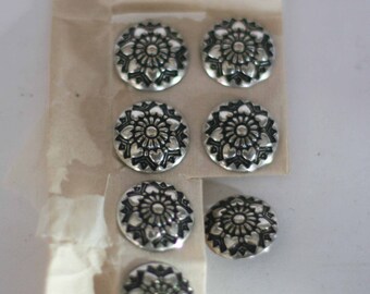 May include: A package of six silver-colored metal buttons with a black floral design. The buttons are arranged in two rows of three.